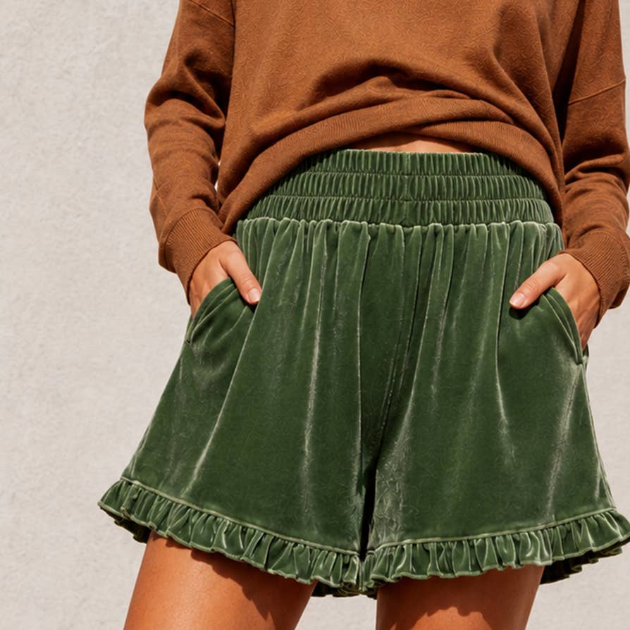 A person wearing dark green velvet shorts with a ruffled hem and side pockets, paired with a brown long-sleeve top, standing against a light gray textured wall.