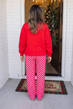 A woman viewed from behind wearing a bright red sweatshirt and red-and-white checkered pajama pants, standing on a porch in front of a decorated Christmas tree and open door.