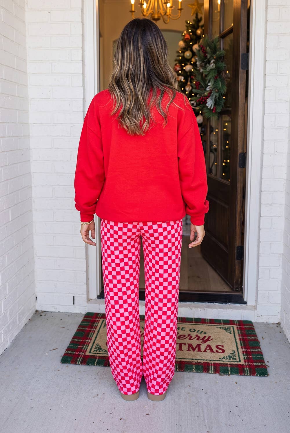 A woman viewed from behind wearing a bright red sweatshirt and red-and-white checkered pajama pants, standing on a porch in front of a decorated Christmas tree and open door.
