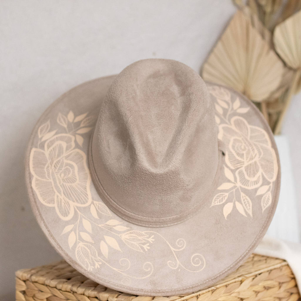 A taupe suede wide-brim hat with artisan painted beige floral print on the brim, resting on a woven straw basket against a neutral background. Crafted in Oaxaca, Mexico.