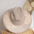 A taupe suede wide-brim hat with artisan painted beige floral print on the brim, resting on a woven straw basket against a neutral background. Crafted in Oaxaca, Mexico.