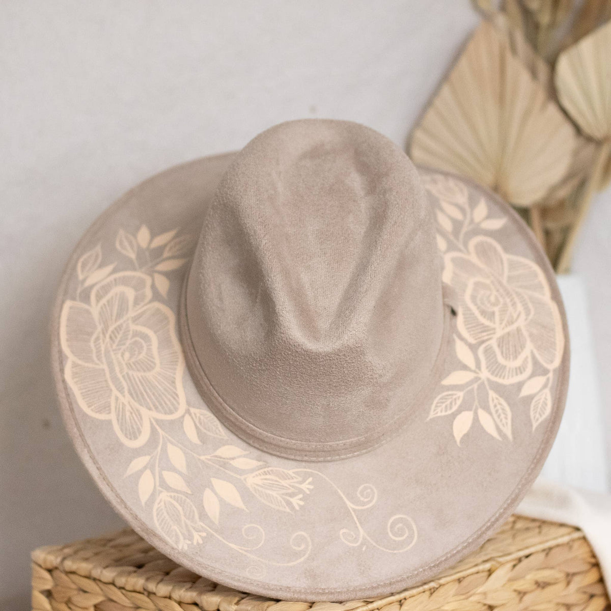A taupe suede wide-brim hat with artisan painted beige floral print on the brim, resting on a woven straw basket against a neutral background. Crafted in Oaxaca, Mexico.