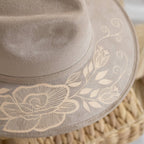 Close-up of a taupe suede hat with an artisan painted beige floral and scroll vine embroidery design on the brim, resting on a woven straw surface. Crafted in Oaxaca, Mexico.