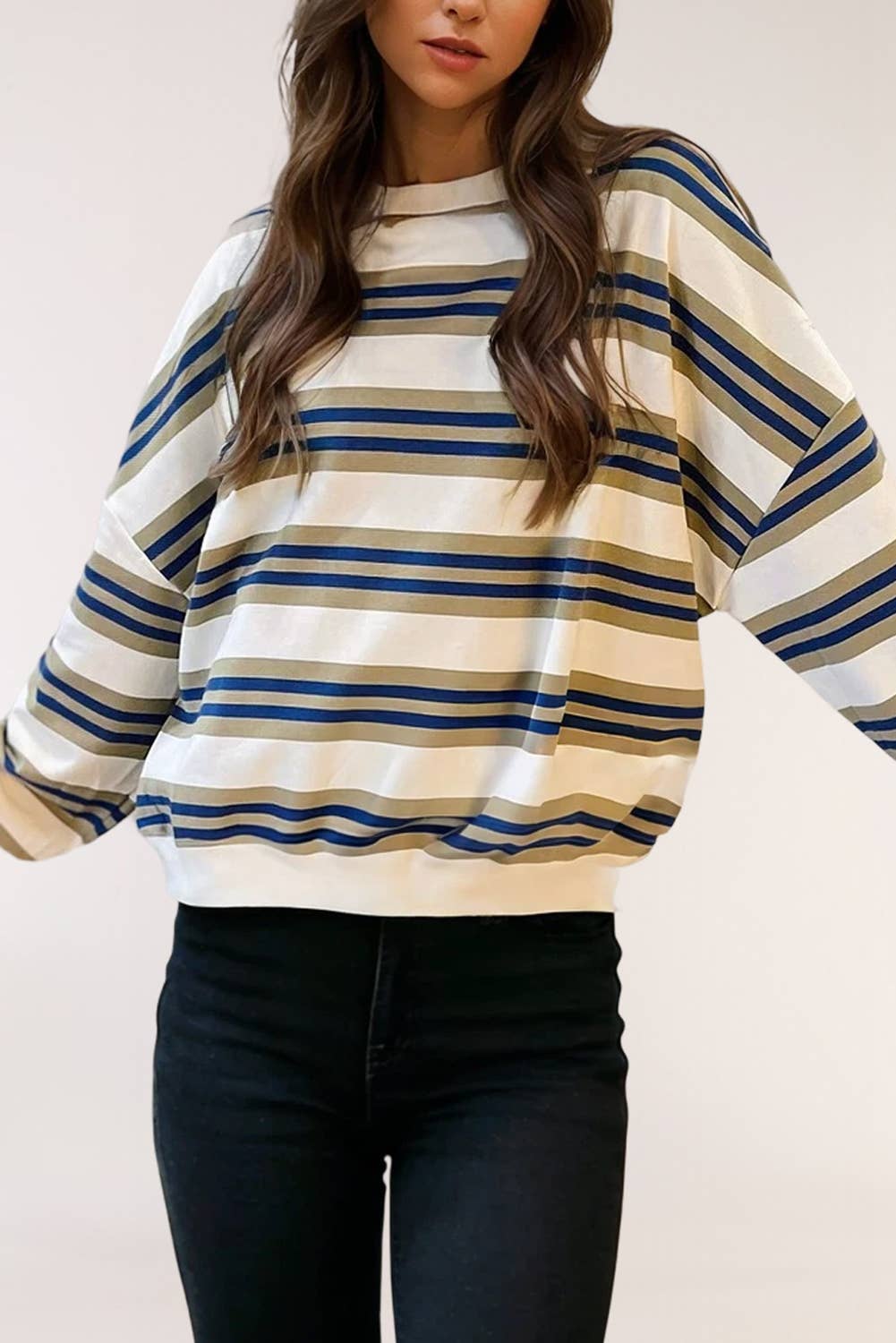 A woman wearing a loose-fit crewneck sweatshirt with horizontal stripes in navy blue, olive green, and cream, paired with black jeans.