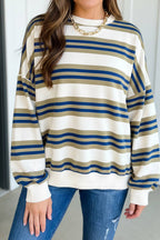 A woman wearing a long-sleeve crewneck sweatshirt with horizontal stripes in cream, navy blue, and tan, paired with blue jeans and a gold chain necklace.