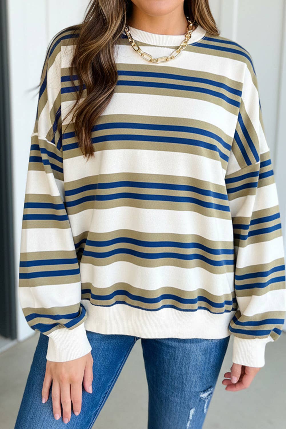 A woman wearing a long-sleeve crewneck sweatshirt with horizontal stripes in cream, navy blue, and tan, paired with blue jeans and a gold chain necklace.