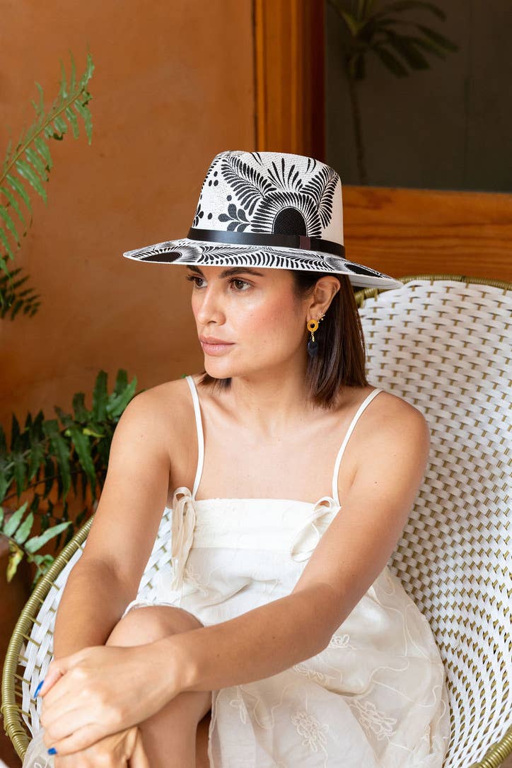 A woman wearing a white fedora-style hat with an artisan painted black botanical pattern and a black band, photographed indoors in a softly blurred room. Crafted in Oaxaca, Mexico.