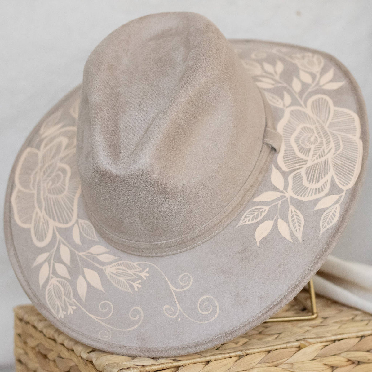 A light taupe suede wide-brim hat with an artisan painted cream floral embroidery on the brim, displayed on a woven straw basket. Crafted in Oaxaca, Mexico.