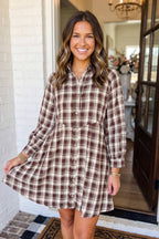A woman modeling a brown and beige plaid button-up shirt dress with long sleeves, standing in a doorway.