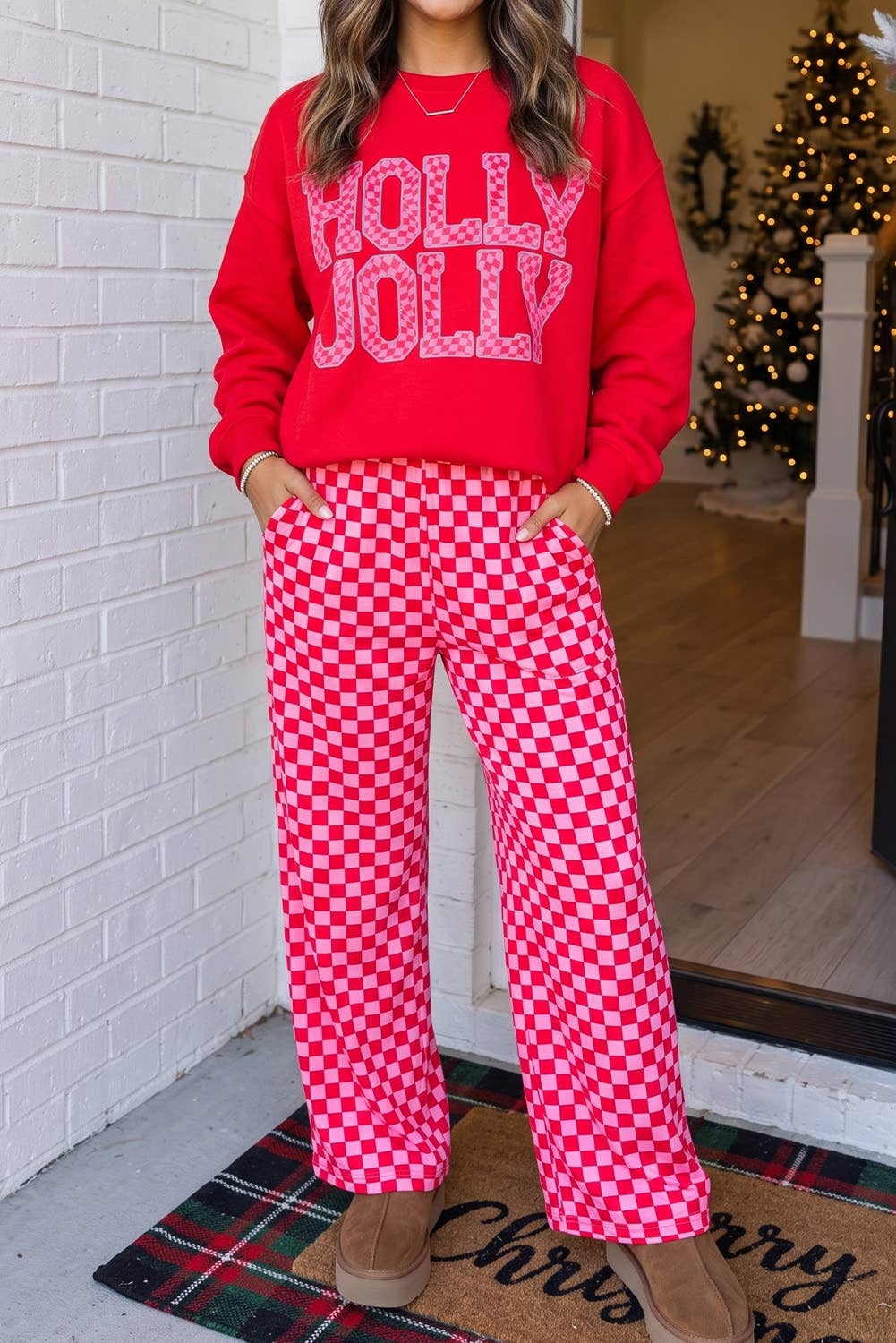 A woman wearing a festive red sweatshirt with 'HOLLY JOLLY' in pink lettering and matching red-and-white checkered pants, standing in a doorway with a Christmas tree in the background.
