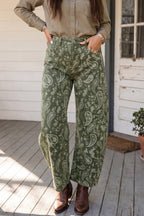 A person wearing barrel wide-leg sage green pants with a light paisley pattern, paired with a beige button-up shirt and brown ankle boots, standing on a wooden porch.