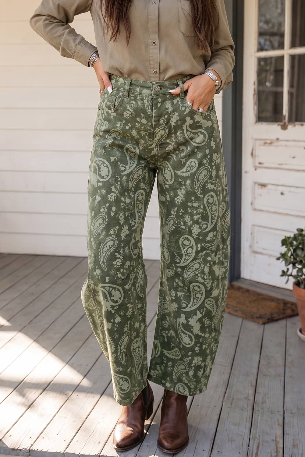 A person wearing barrel wide-leg sage green pants with a light paisley pattern, paired with a beige button-up shirt and brown ankle boots, standing on a wooden porch.
