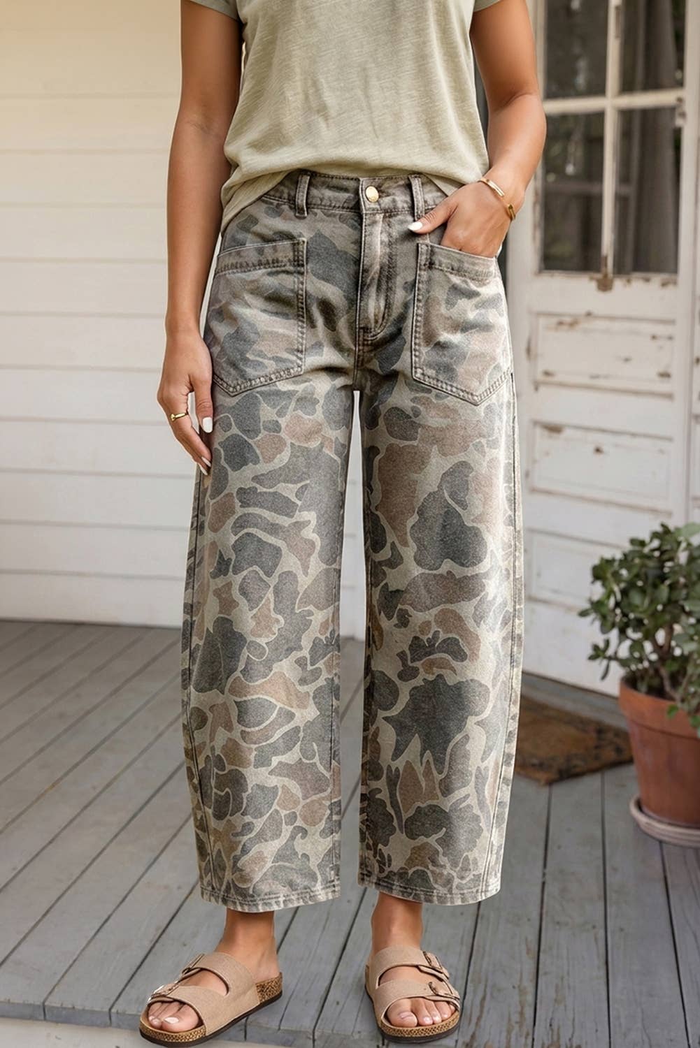 A person wearing wide-leg cropped jeans with a muted camouflage pattern in gray, beige, and brown, paired with a light olive top and tan sandals on a porch.