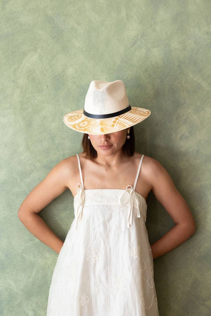 Woman wearing a white dress and an artisan painted sun hat against a green wall. Crafted in Oaxaca, Mexico.