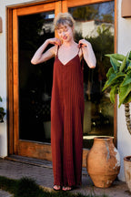 A woman wearing a rust-brown pleated maxi dress with thin spaghetti straps, standing in front of a glass door with wooden frames. She is holding the straps of the dress, and a large terracotta pot and green plant are visible on either side.