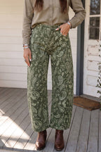 A person wearing barrel wide-leg sage green pants with a beige paisley pattern, styled with a khaki button-down shirt and brown leather ankle boots, standing on a wooden porch.