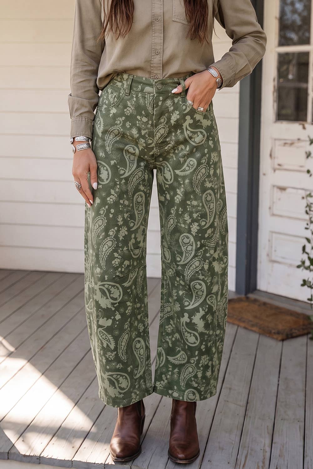 A person wearing barrel wide-leg sage green pants with a beige paisley pattern, styled with a khaki button-down shirt and brown leather ankle boots, standing on a wooden porch.