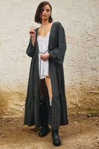 A woman stands outdoors against a textured beige wall wearing a long black open-front cardigan with a white geometric pattern over a white camisole, brown leggings, and black boots.