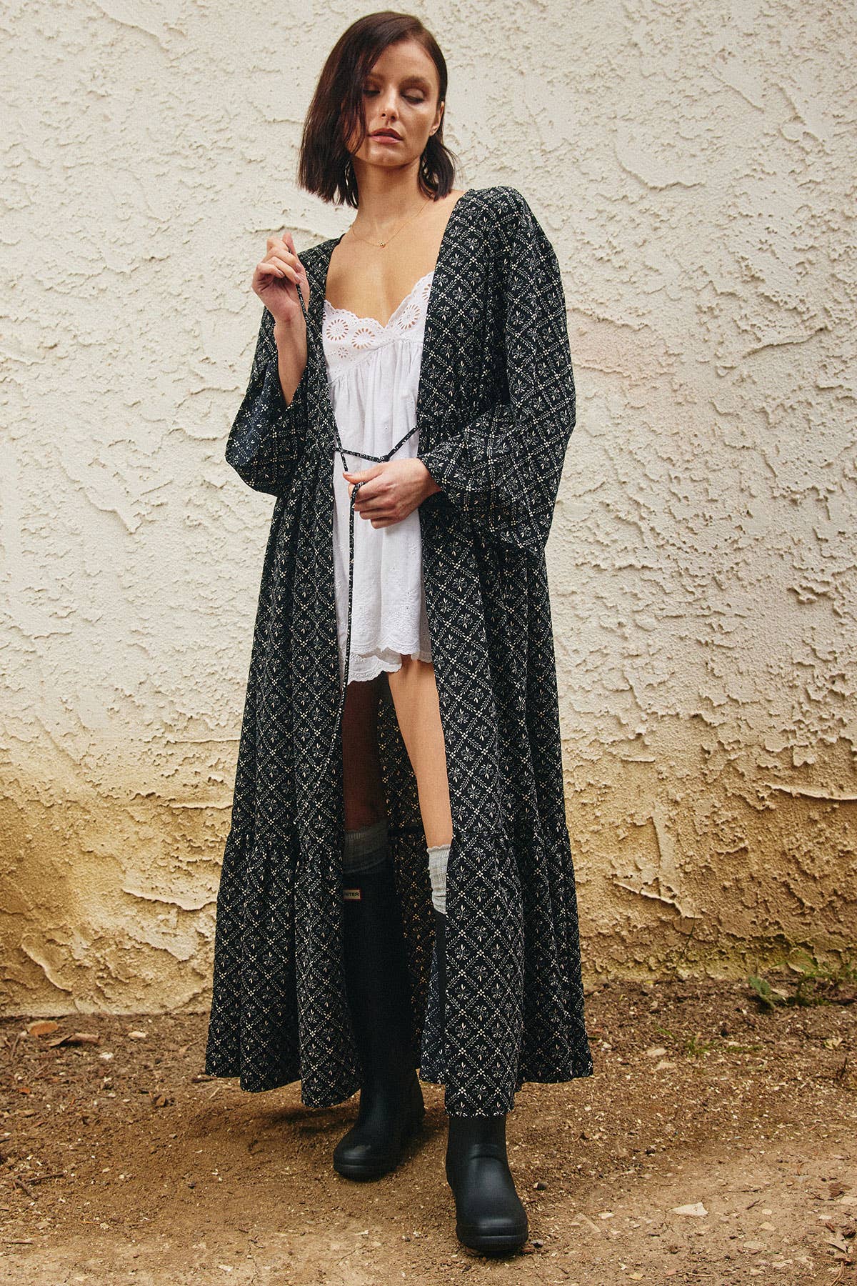 A woman stands outdoors against a textured beige wall wearing a long black open-front cardigan with a white geometric pattern over a white camisole, brown leggings, and black boots.
