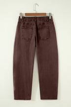 A pair of dark brown cropped jeans with an elastic waistband and back pockets, hanging on a wooden hanger against a plain off-white wall.