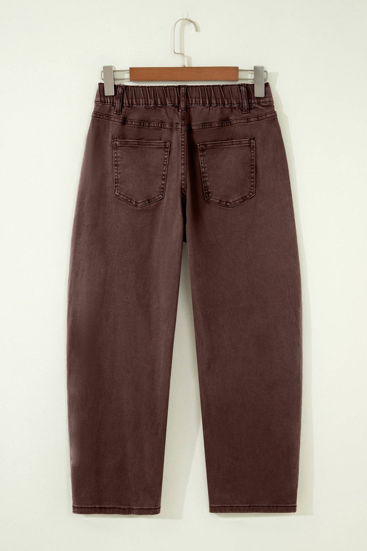 A pair of dark brown cropped jeans with an elastic waistband and back pockets, hanging on a wooden hanger against a plain off-white wall.