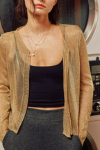 A woman wearing a shimmering metallic gold cardigan over a black crop top and gray skirt, standing in front of a washing machine.